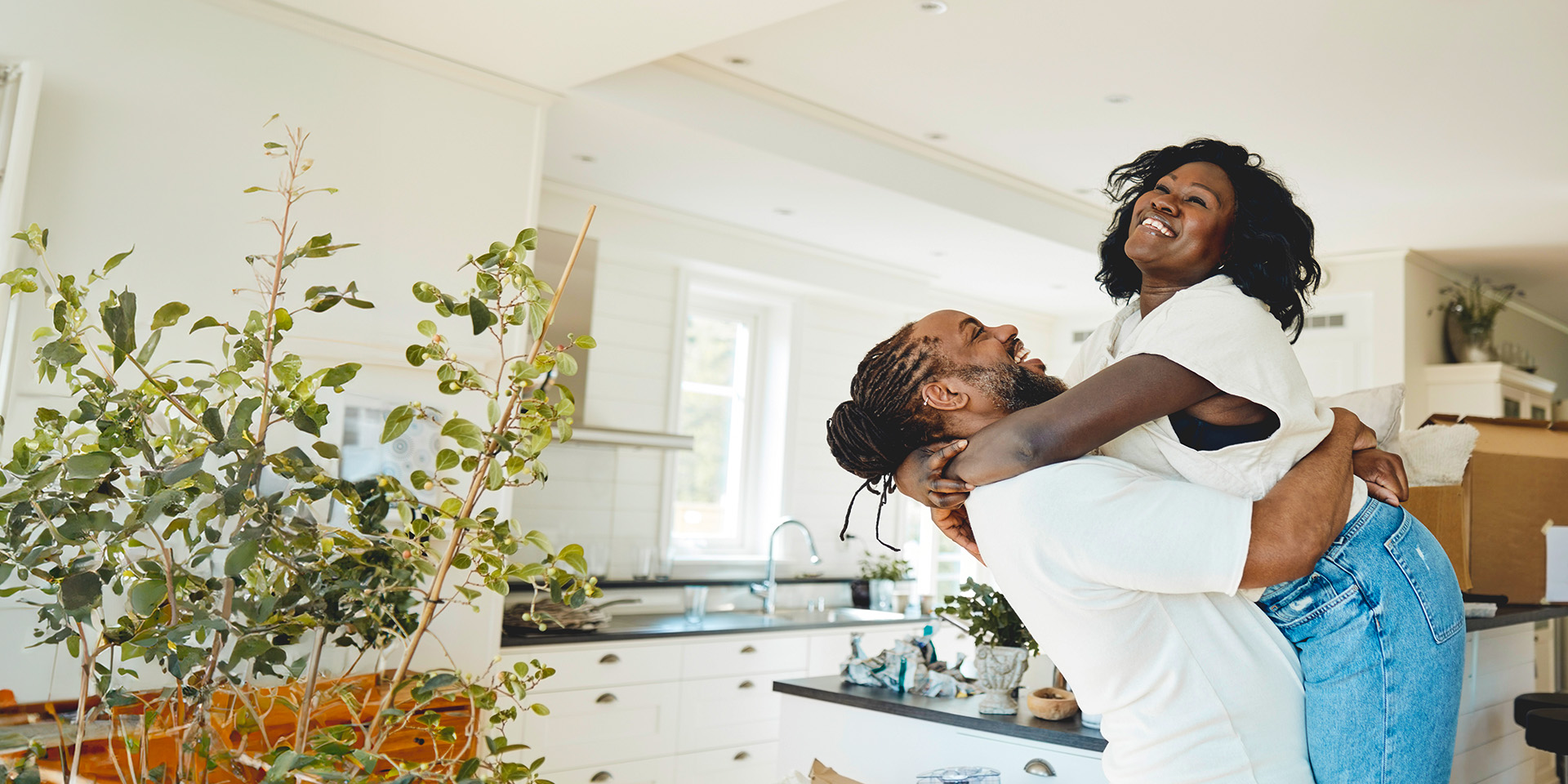 New homeowners celebrate in their kitchen, surrounded by unpacked boxes. Husband joyfully lifts his wife in a warm embrace, marking a moment of happiness in their first home together.