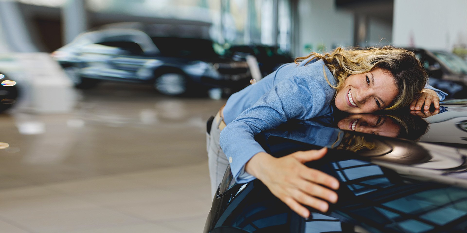 An excited woman hugging the front of her new car inside a car dealership.