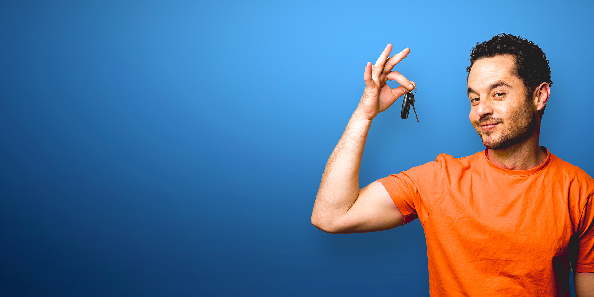 A smirking man dangling car keys up by his head in front of a blue background.