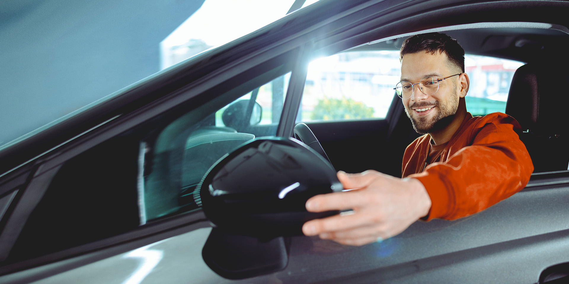 A new graduate sitting in the driver's seat of his car and fixing the side mirror through the window.