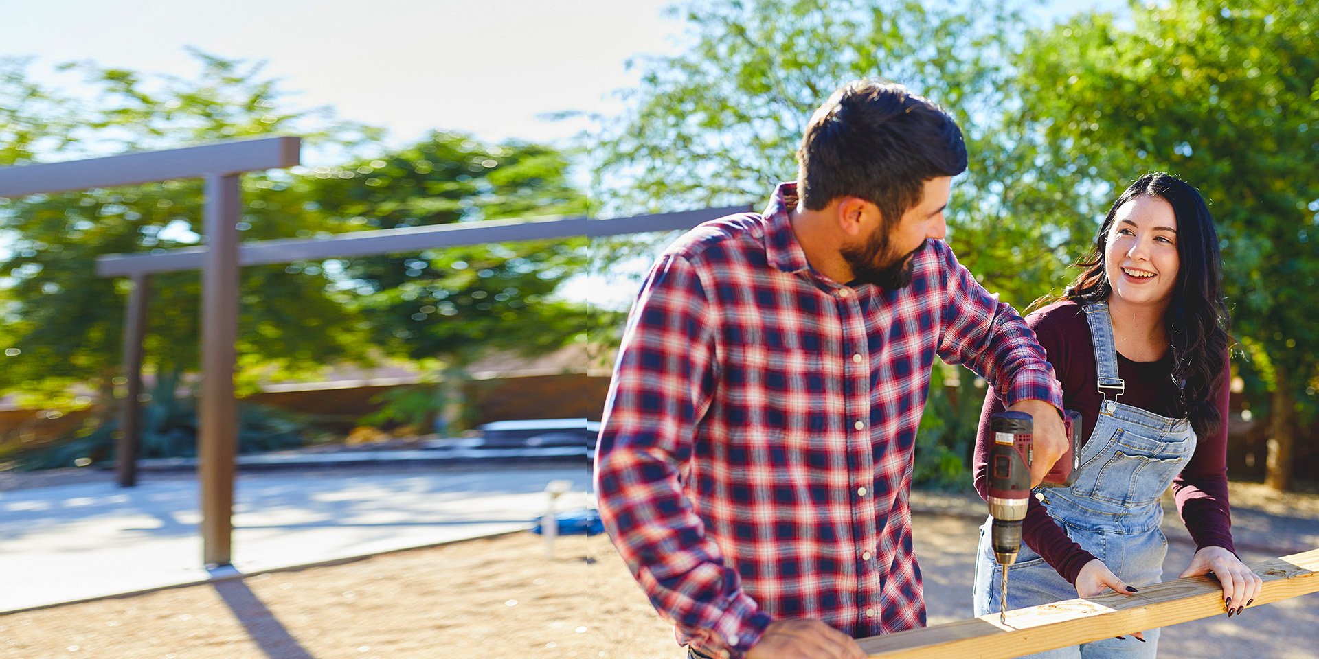 A couple collaborates in their backyard project: the man drills into plywood as the woman provides steady support, building together with enthusiasm.