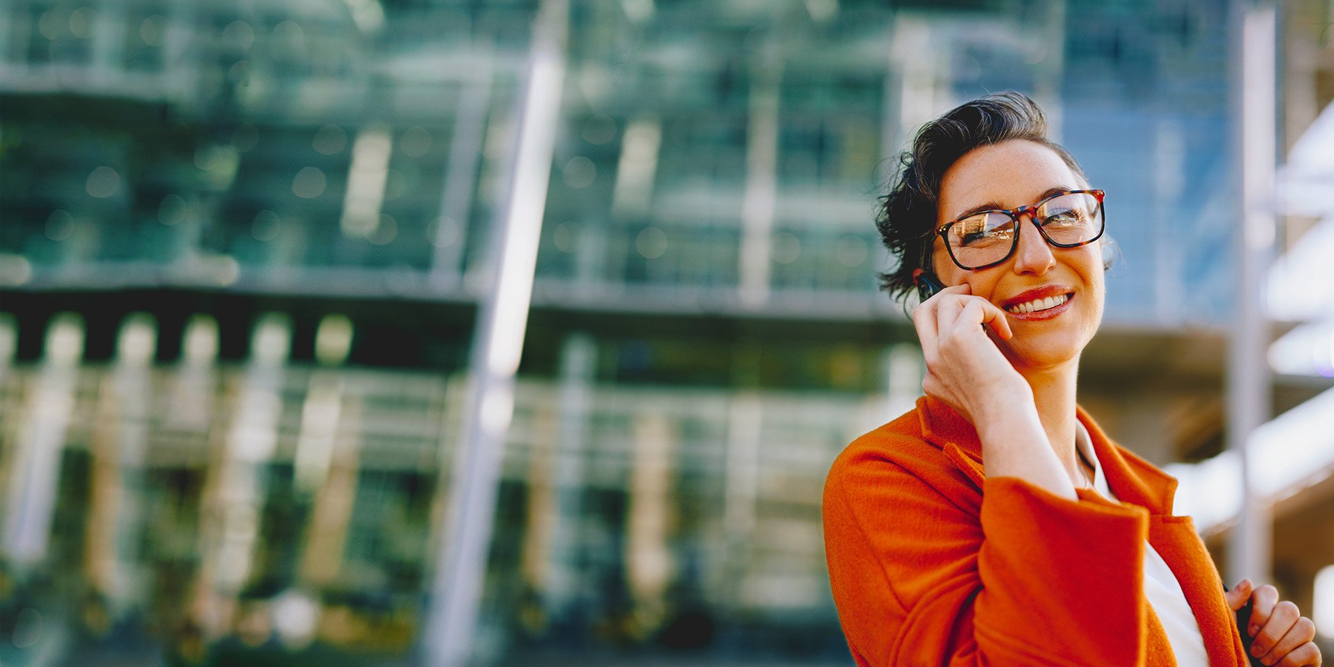 A happy businesswoman with glasses who is on the phone outside.