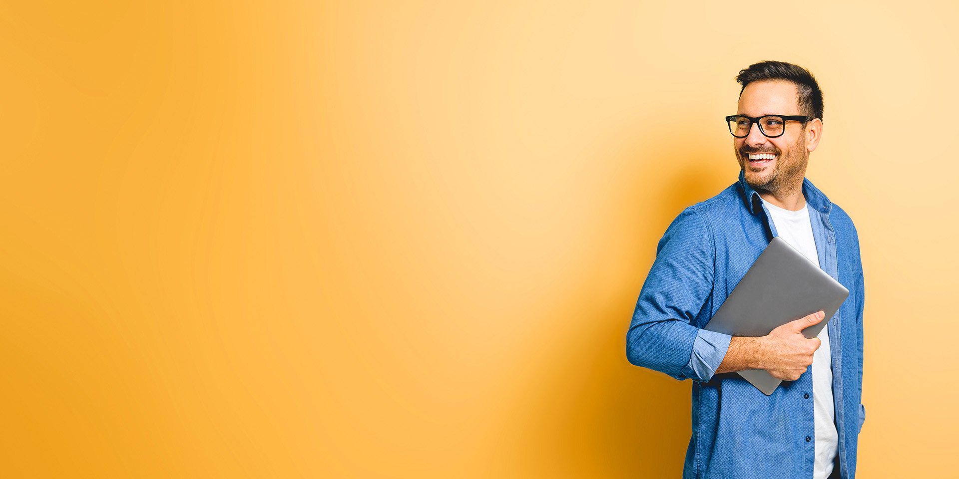 A joyful man with glasses holds a laptop to his chest with one hand, smiling over his shoulder against an orange background.