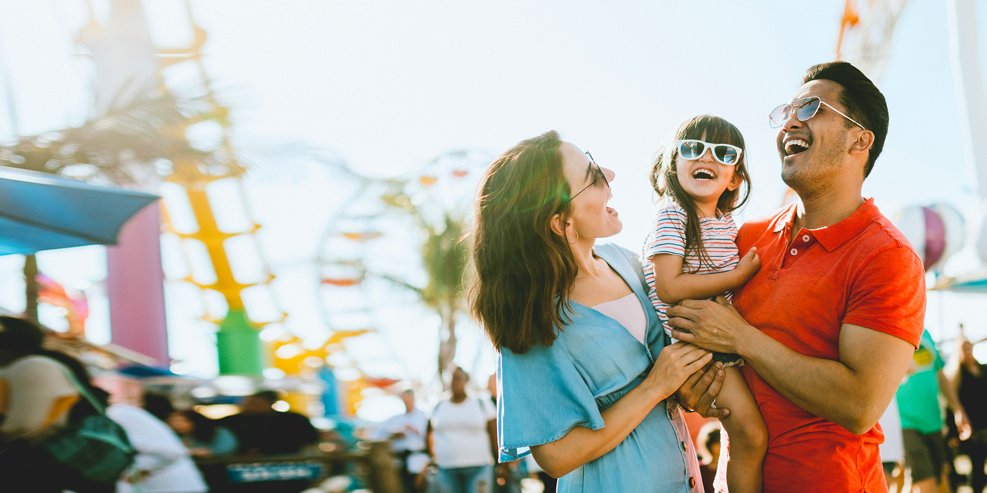 Two parents savoring a day of fun at a theme park with their young daughter, all wearing sunglasses. The father lovingly holds the girl while the mother embraces them both.