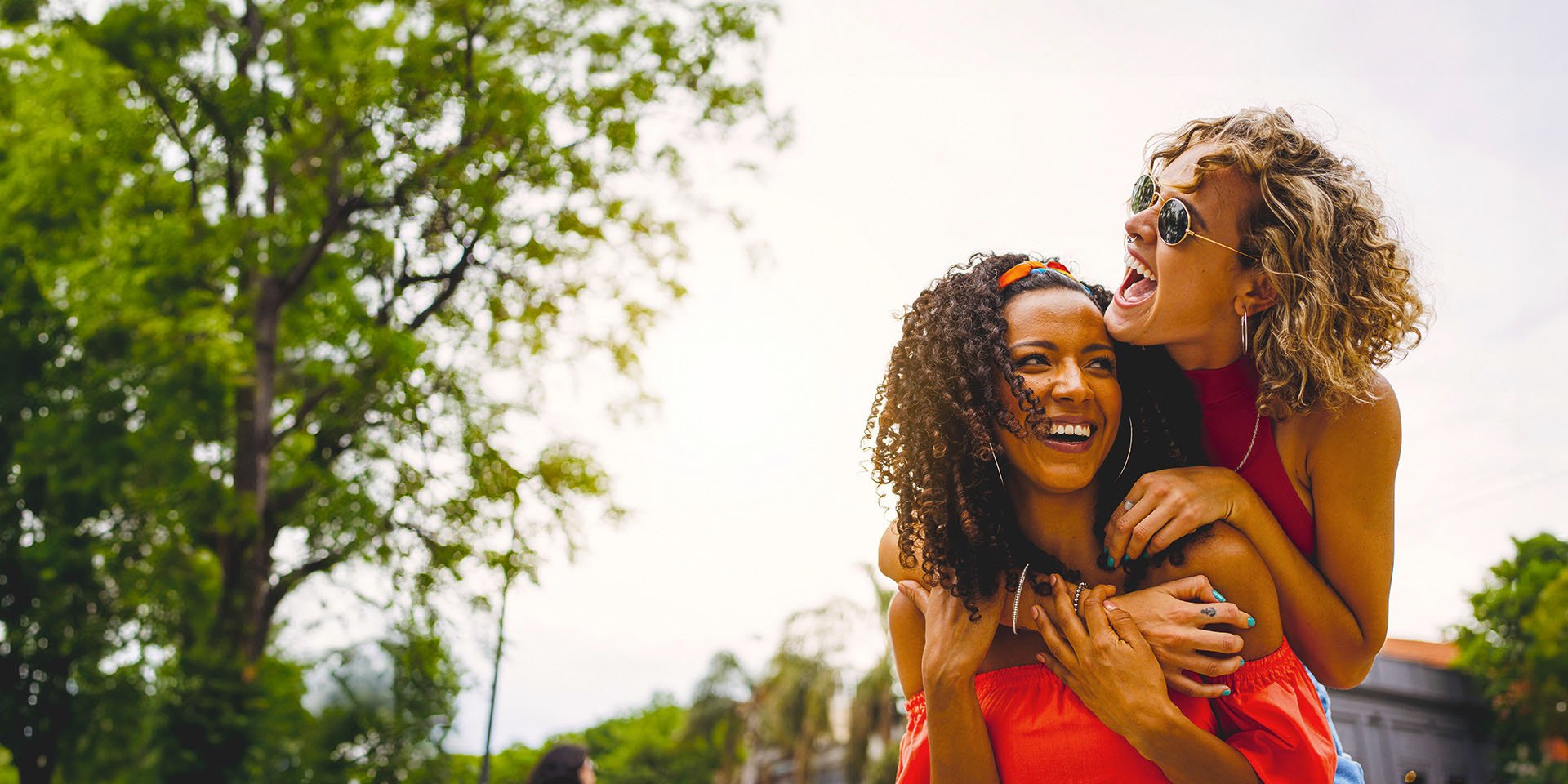 Two joyful women having a great time in an outdoor park. One woman is on the other's back, and both are sharing laughter.