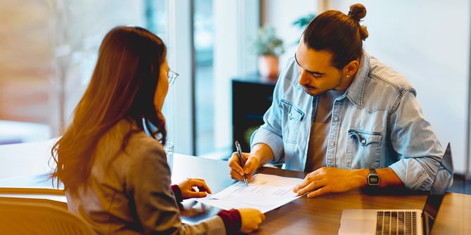 A bank teller helping a young man fill out a written form over the counter.