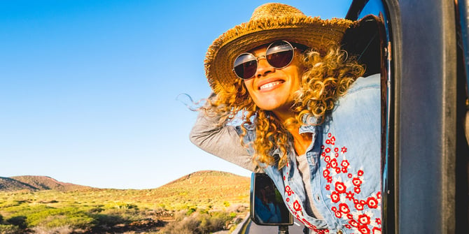 A happy woman in a straw hat and sunglasses hanging out of a car window driving down a paved road in the desert.