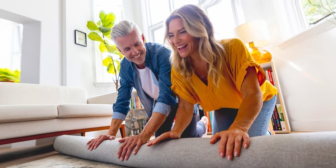 A couple on the floor of their living room excitedly rolling out a new rug on the floor together.