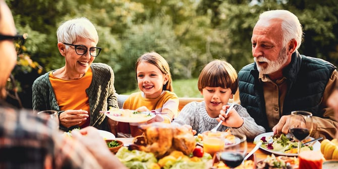 Grandparents sitting on a bench on either side of their granddaughter and grandson while enjoying an outdoor family dinner.